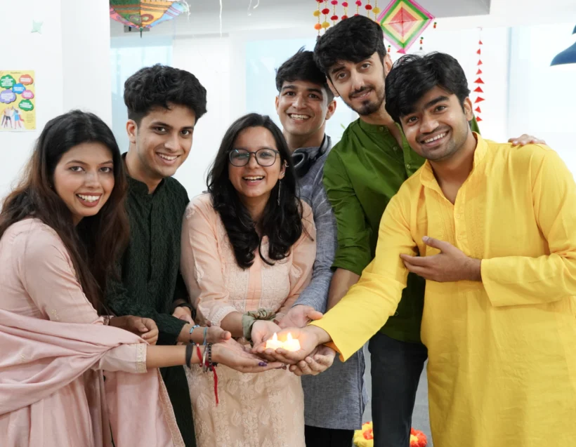 Six people in traditional Indian clothing stand indoors, smiling and holding a lit clay lamp together, with festive decorations visible in the background.