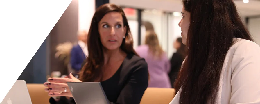 Two women sitting at a table with laptops, engaged in conversation in an office setting.