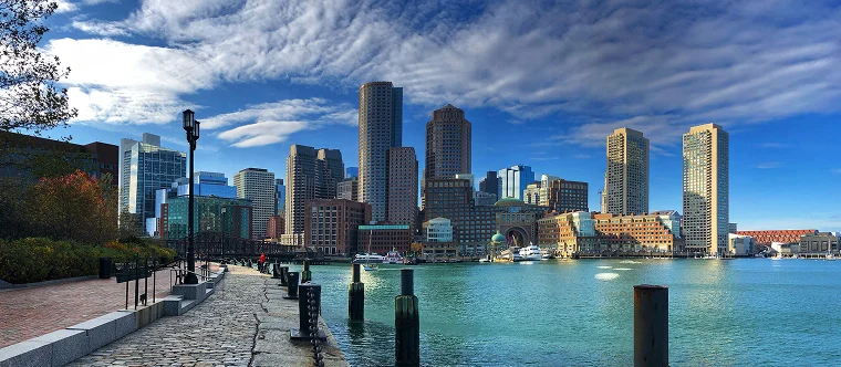 Skyline view of downtown Boston with modern high-rise buildings, waterfront, and a partly cloudy sky, seen from a cobblestone walkway along the harbor.
