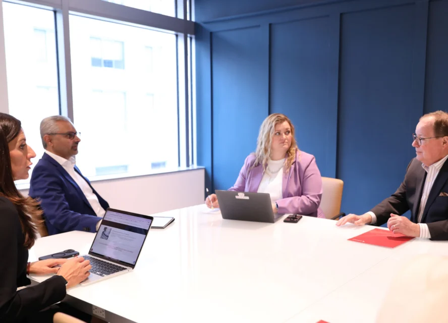 Four people in business attire sit around a conference table with laptops and documents, engaged in a meeting in a modern office.