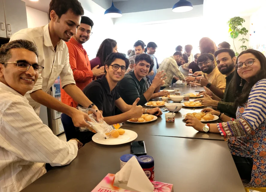 A group of people sit and stand around a table, serving and enjoying snacks, smiling and interacting in a brightly lit, informal setting.