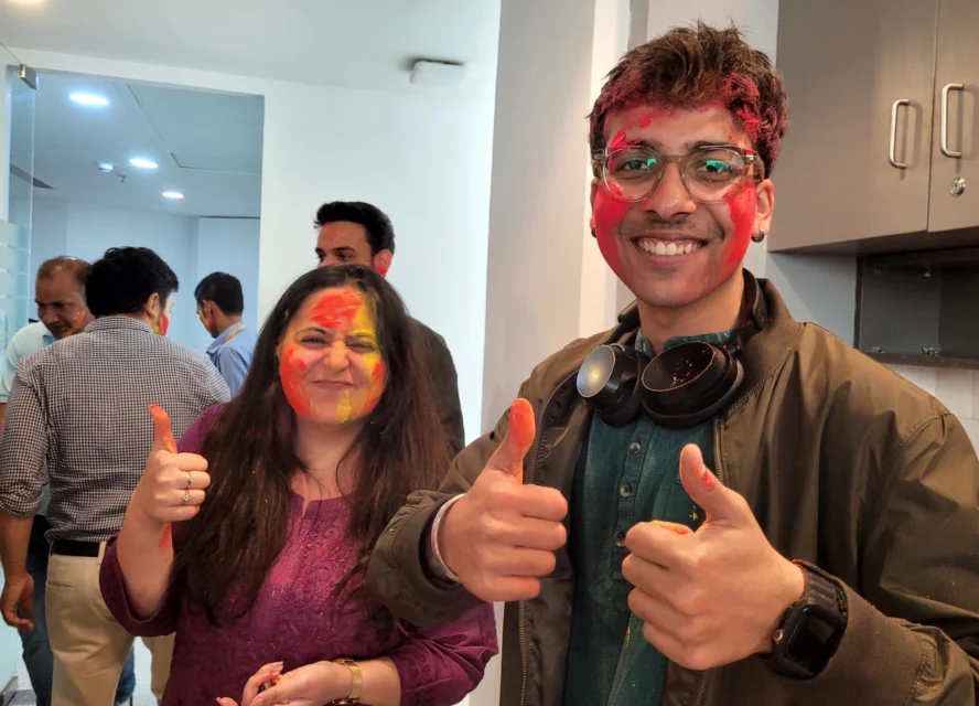 Two people with colorful powder on their faces smile and give thumbs up in an indoor setting; others are visible in the background.