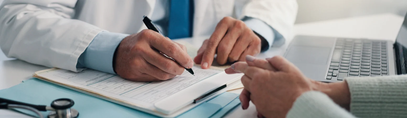 A doctor in a white coat writes on a medical form while talking to a patient across the desk; a stethoscope and laptop are nearby.
