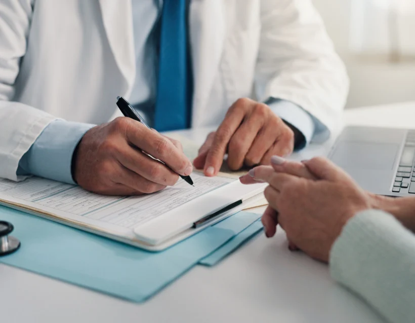 A doctor in a white coat writes on a medical form while talking to a patient across the desk; a stethoscope and laptop are nearby.