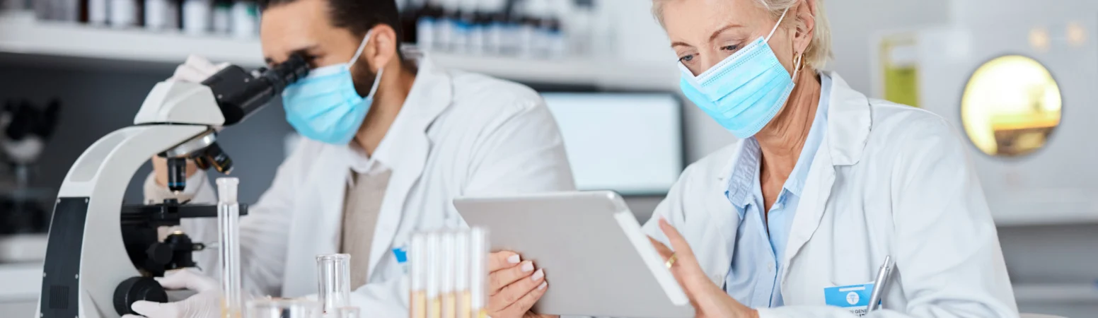 Two scientists in lab coats and face masks work in a laboratory; one uses a microscope while the other examines a tablet. Test tubes and lab equipment are on the table.