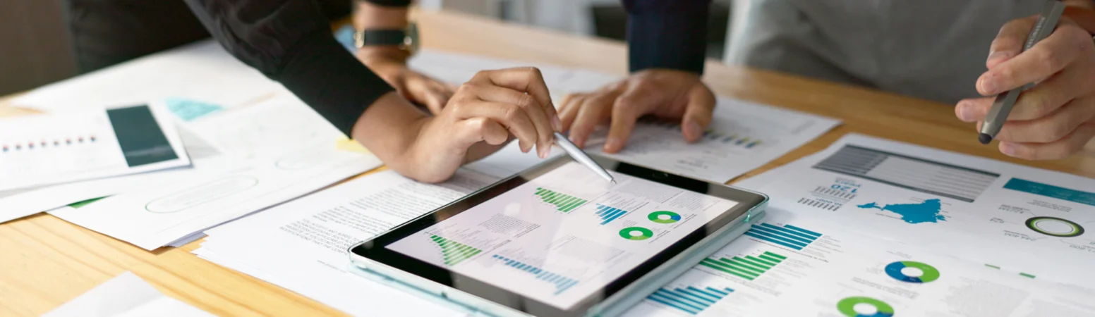 Two people review graphs and charts on printed documents and a tablet, analyzing data together at a desk.