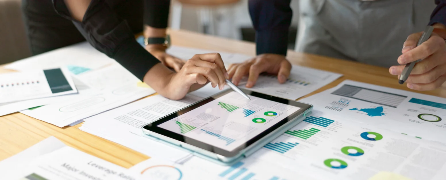 Two people review graphs and charts on printed documents and a tablet, analyzing data together at a desk.
