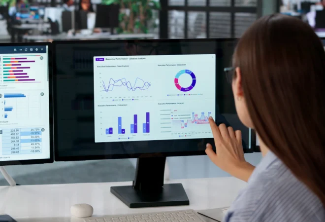 A woman reviews charts and graphs on dual monitors at an office desk, analyzing data displayed in bar, line, and pie chart formats.