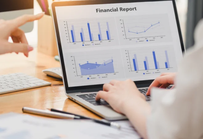 Two people review a laptop displaying a financial report with bar and line charts. One person points at the screen, and documents are spread on the desk.
