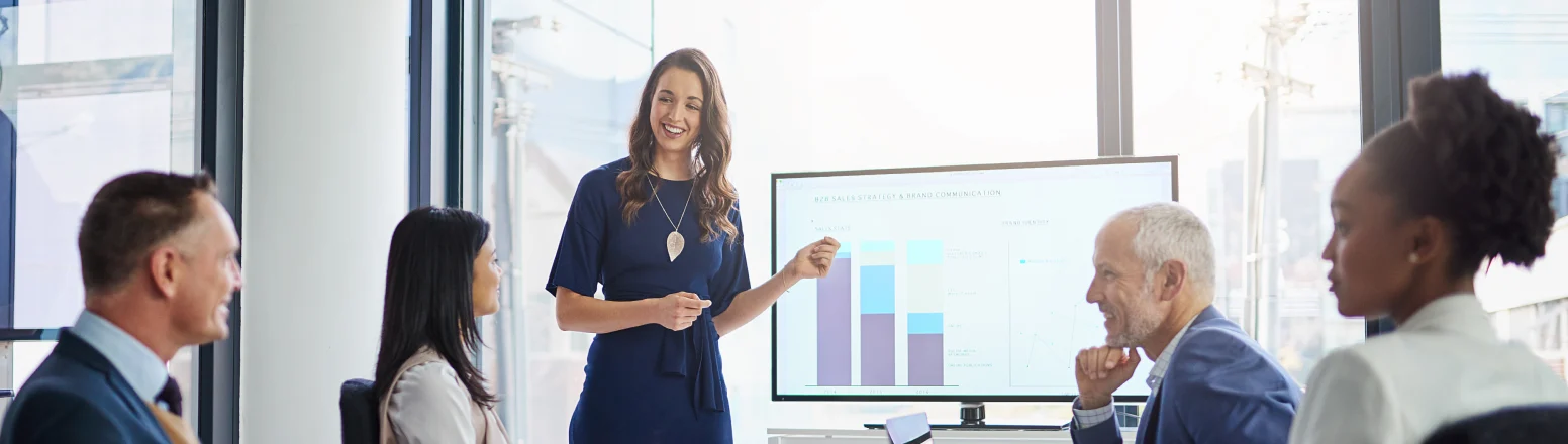 A woman stands and presents a bar and line graph on a screen to four colleagues seated around a table in a modern office meeting room.
