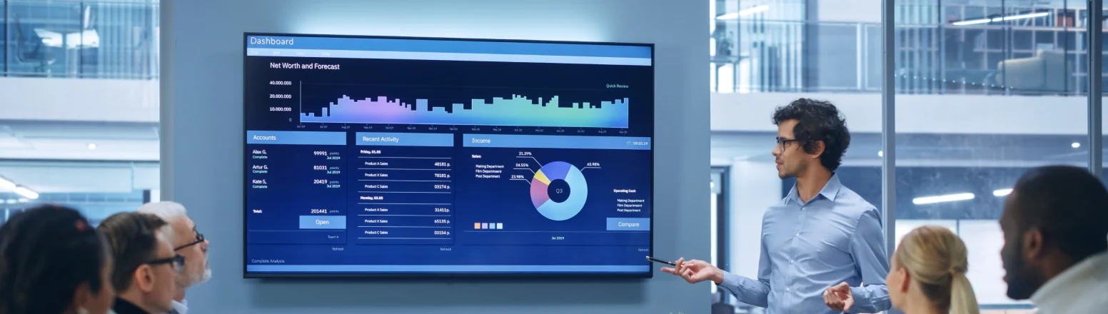 A man in business attire presents financial data on a large screen to a group of colleagues in a modern office meeting room.