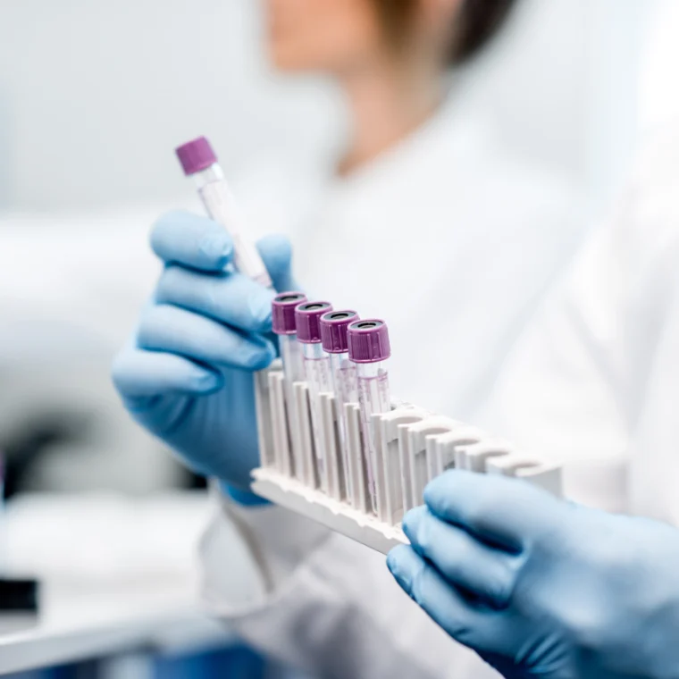 Gloved hands holding test tubes with purple caps, placing them into a white rack in a laboratory setting.