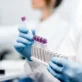 Gloved hands holding test tubes with purple caps, placing them into a white rack in a laboratory setting.