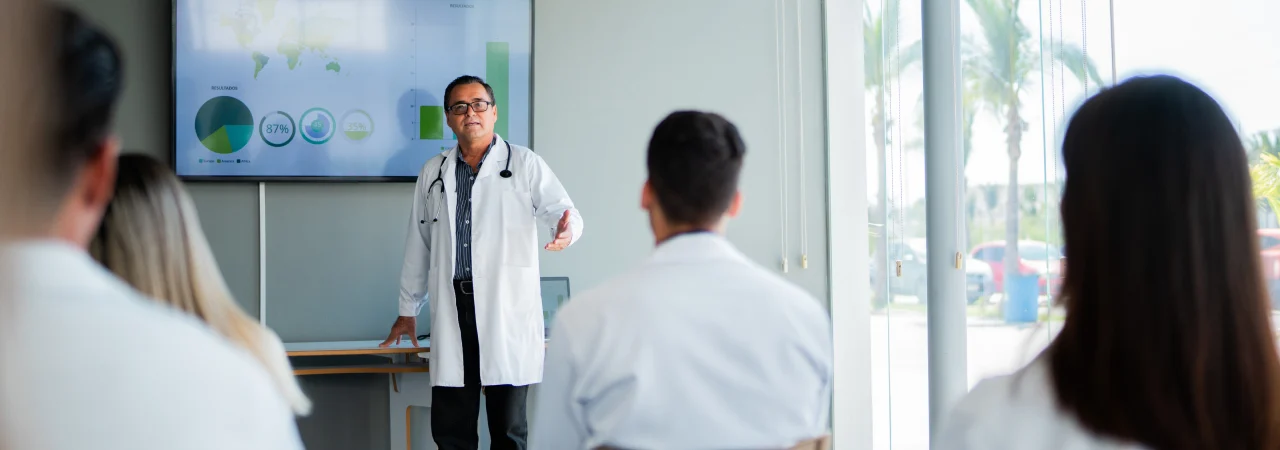 A doctor gives a presentation with charts displayed on a screen to a group of people in lab coats seated in a conference room with large windows.