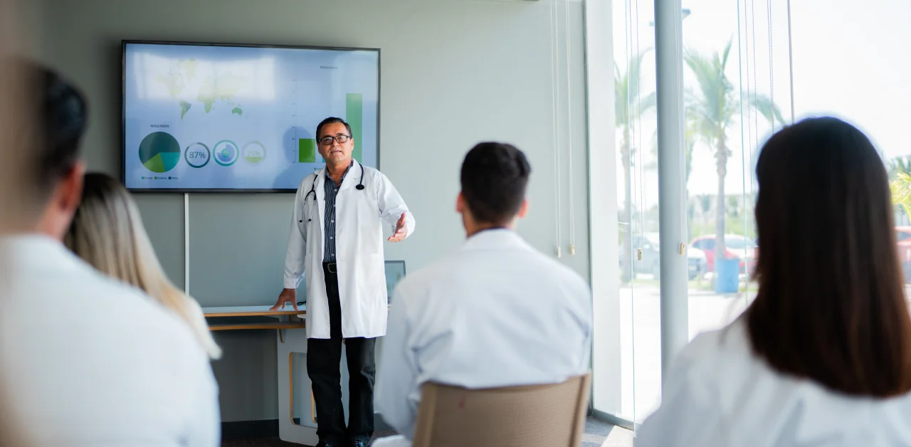 A doctor gives a presentation with charts displayed on a screen to a group of people in lab coats seated in a conference room with large windows.