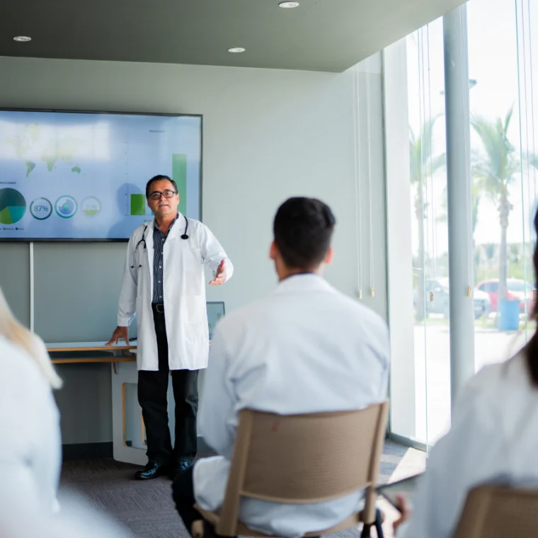 A doctor gives a presentation with charts displayed on a screen to a group of people in lab coats seated in a conference room with large windows.