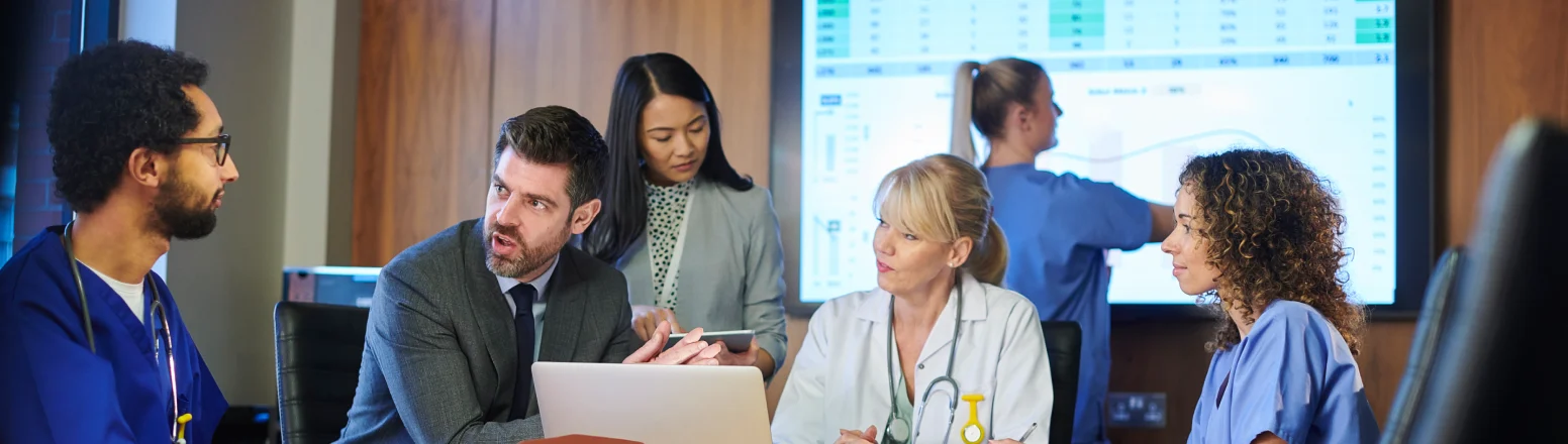 A group of healthcare professionals and a man in a suit have a meeting around a table, with charts displayed on a screen in the background.
