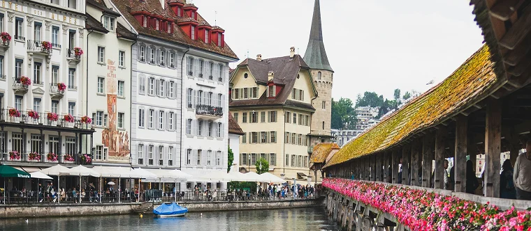 A historic wooden bridge decorated with flowers crosses a river beside old European buildings, with people dining outdoors and a boat on the water.