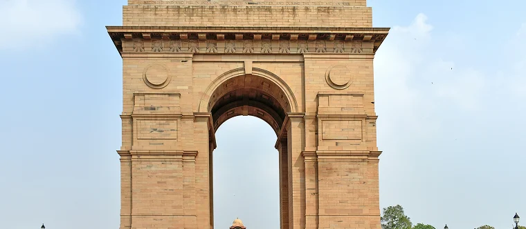 The image shows India Gate, a large stone war memorial arch located in New Delhi, India, under a clear sky.