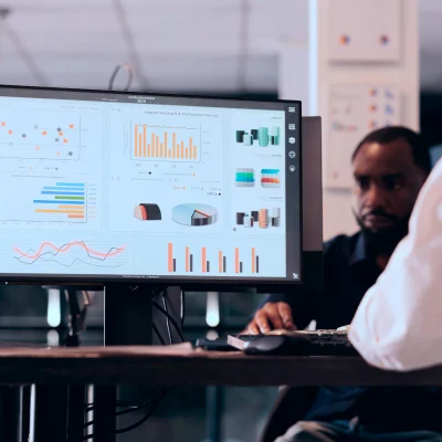 A computer monitor displays various data charts and graphs while a person works at a desk in an office setting.