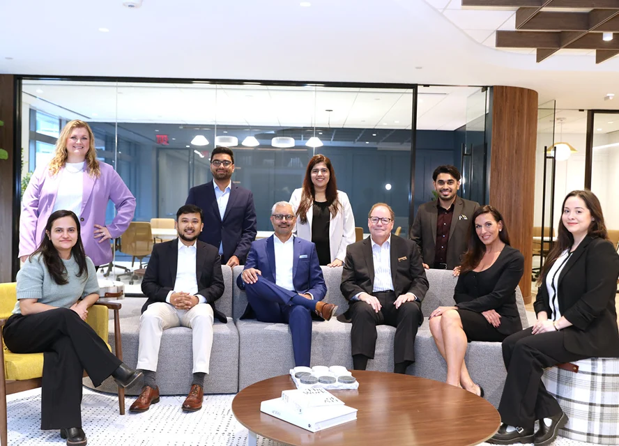 A group of eleven professionally dressed people pose for a photo in a modern office lounge, with some seated on a sofa and others standing behind.