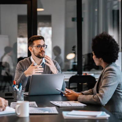 Two colleagues sit at a desk in a modern office, engaged in a discussion. One gestures while speaking, the other listens and holds a smartphone. A laptop and documents are on the table.