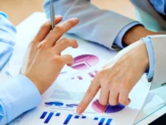 Two people in business attire discuss financial charts and graphs on printed documents at a desk, with a pen and a tablet visible.