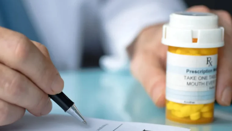 A person writes a prescription on a pad while holding a bottle of yellow pills labeled as prescription medication.