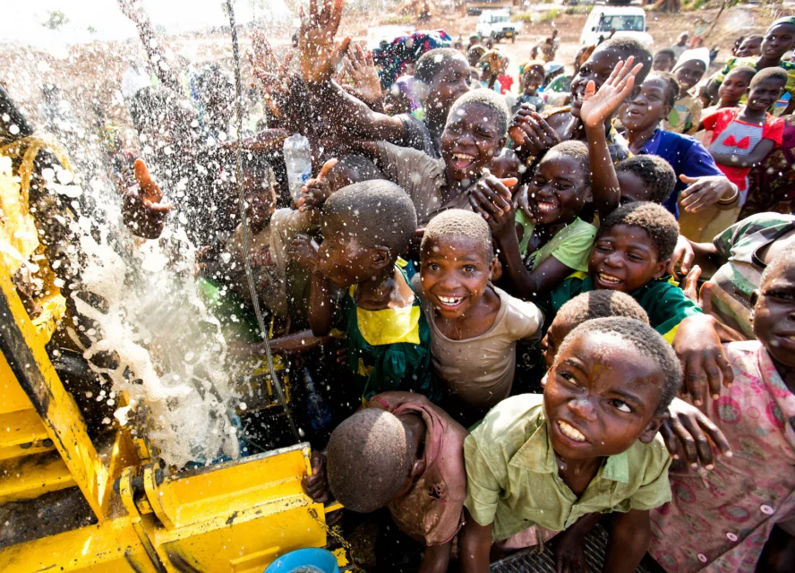 A group of children gather around and smile as water gushes from a newly installed water pump.