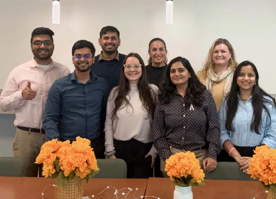 Eight people pose together indoors behind a table decorated with two vases of orange flowers.