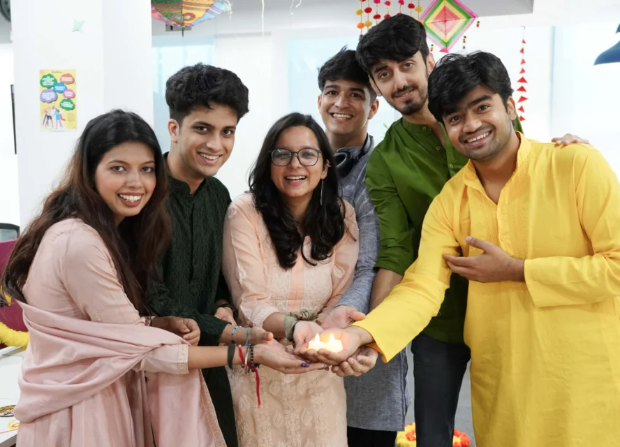 Six people in traditional Indian attire smile and hold a lit diya lamp together, standing indoors with festive decorations in the background.