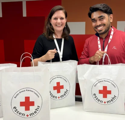 Two people standing behind a table, each holding a white bag labeled "Essen + Mehr" with a red cross logo; more bags are on the table.