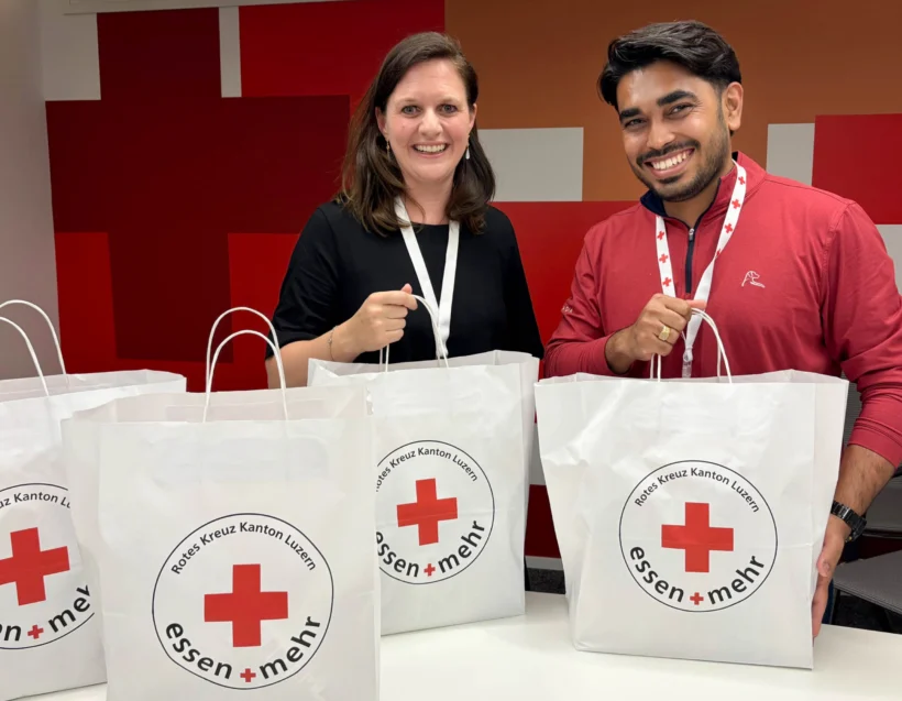 Two people standing behind a table, each holding a white bag labeled "Essen + Mehr" with a red cross logo; more bags are on the table.