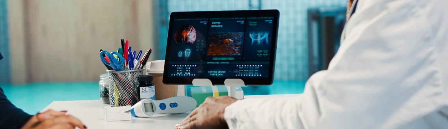 A doctor sits at a desk using a computer displaying medical images and data while talking to a patient whose hands are visible.