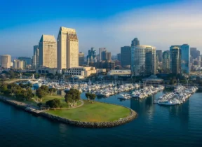 Aerial view of downtown San Diego with high-rise buildings, a marina filled with boats, and a green park peninsula extending into the harbor.