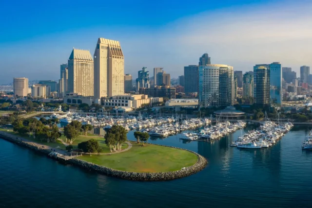 Aerial view of downtown San Diego with high-rise buildings, a marina filled with boats, and a green park peninsula extending into the harbor.