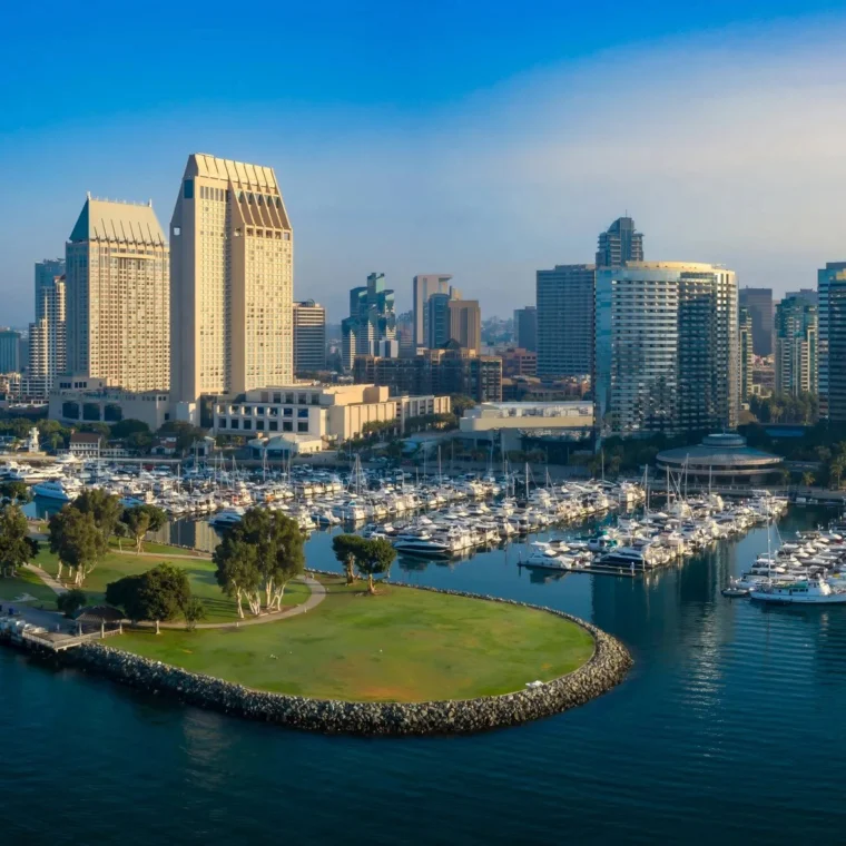 Aerial view of downtown San Diego with high-rise buildings, a marina filled with boats, and a green park peninsula extending into the harbor.