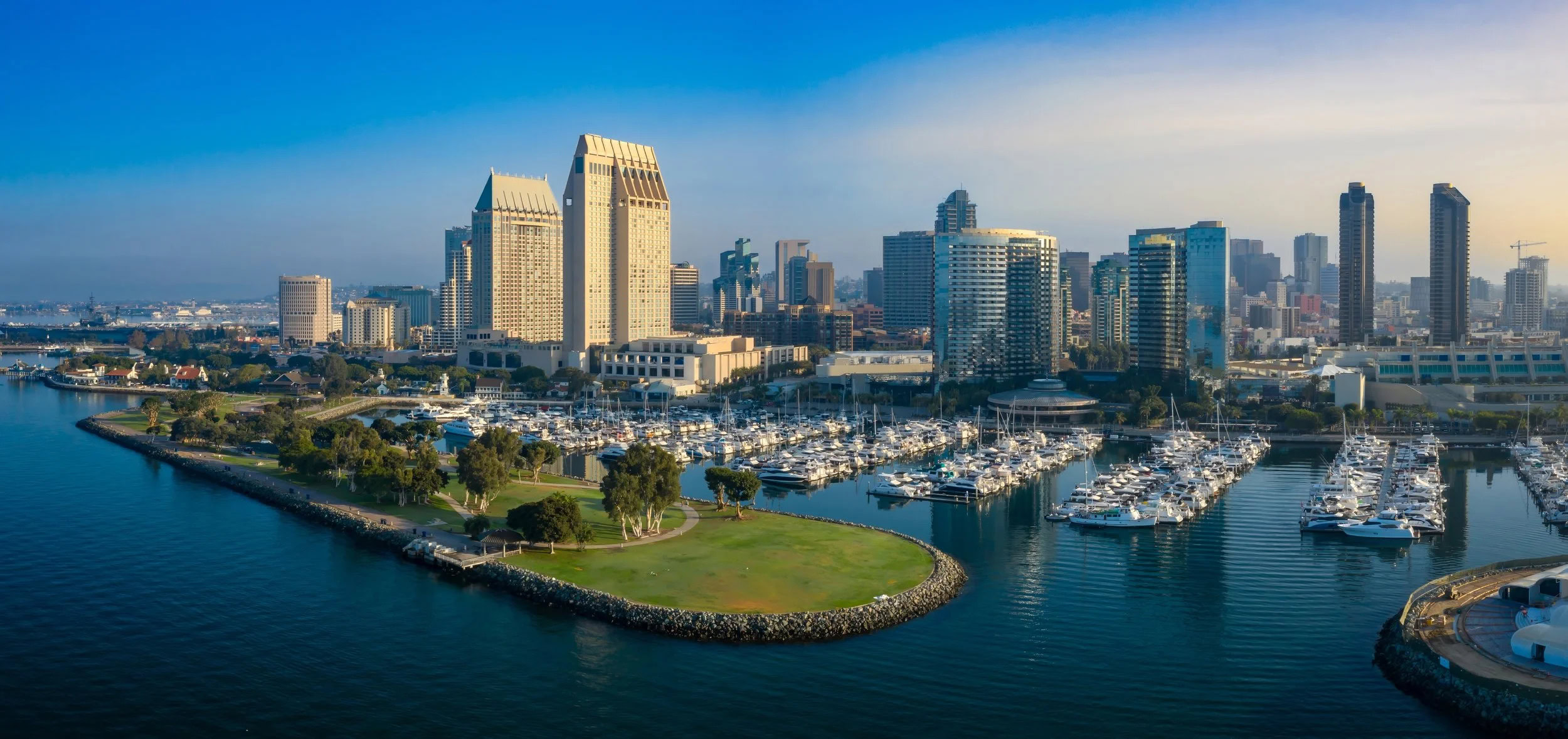 Aerial view of downtown San Diego with high-rise buildings, a marina filled with boats, and a green park peninsula extending into the harbor.