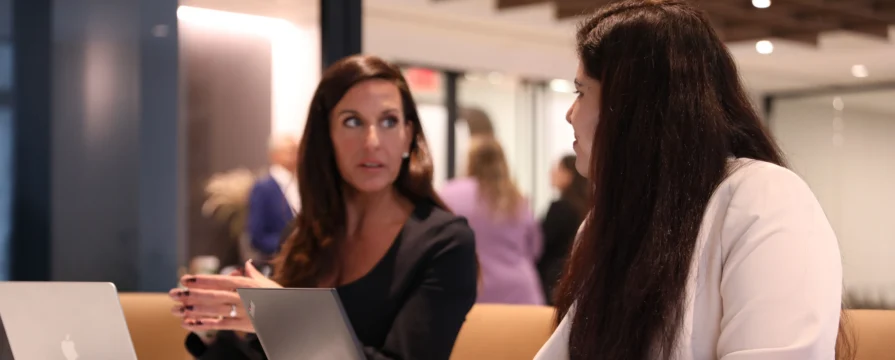 Two women sitting at a table with laptops, engaged in conversation in an office setting.