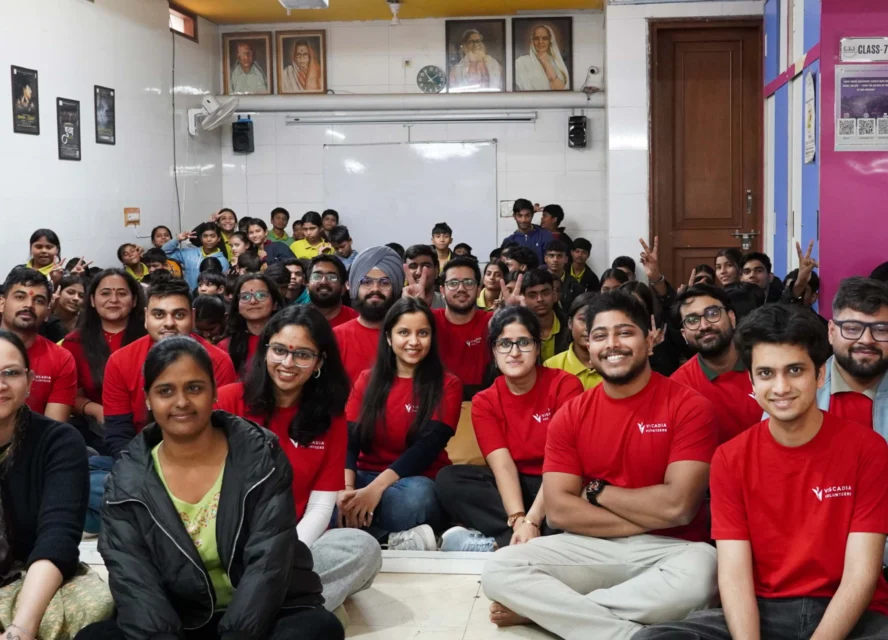 A group of people, some in red shirts, sit and pose for a photo in a classroom with portraits and posters on the wall behind them.